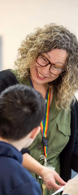 Lecturer with a green shirt and rainbow lanyard smiling while reviewing an art student's work in a classroom with artwork in the background. Lecturer with a green shirt and rainbow lanyard smiling while reviewing an art student's work in a classroom with artwork in the background.